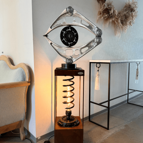 Sur cette image on y voit une sculpture lumineuse réalisée avec des pièces mécaniques recyclées, la partie du haut représente un oeil et est posé sur un socle. A l'intérieur de celui-ci il y a une lumière allumée, elle est réalisée avec d'autres pièces mécaniques recyclées.