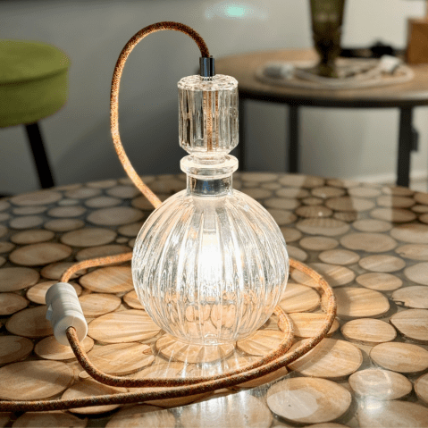 Sur cette photo, l'on voit une carafe en verre de forme ronde posée sur une table. Cette carafe est allumée car elle est transformée en lampe de table, elle a un cordon électrique en tissu dans les tons orange.