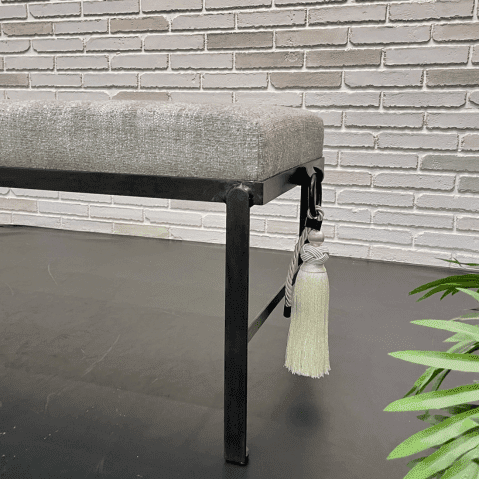 Cette image montre une partie d'une banquette faite en structure métallique noir, une assise en tissu blanc crème légèrement chiné et une décoration sur le côté qui est un anneau en métal noir où pend un pompon à frange comme les attaches de rideau couleur crème.