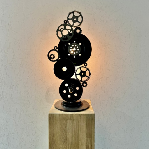 Sculpture Bon Débarras ! présentée sur un socle en bois, avec ses engrenages et disques de freins projetant des ombres sur le mur éclairé.